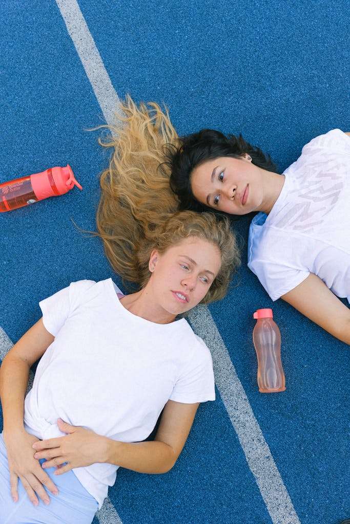 Two women in activewear resting on a track with water bottles, promoting fitness and relaxation.